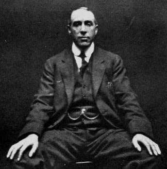 Portrait noir et blanc de Harry Price assis, mains sur les genoux, fond sombre.